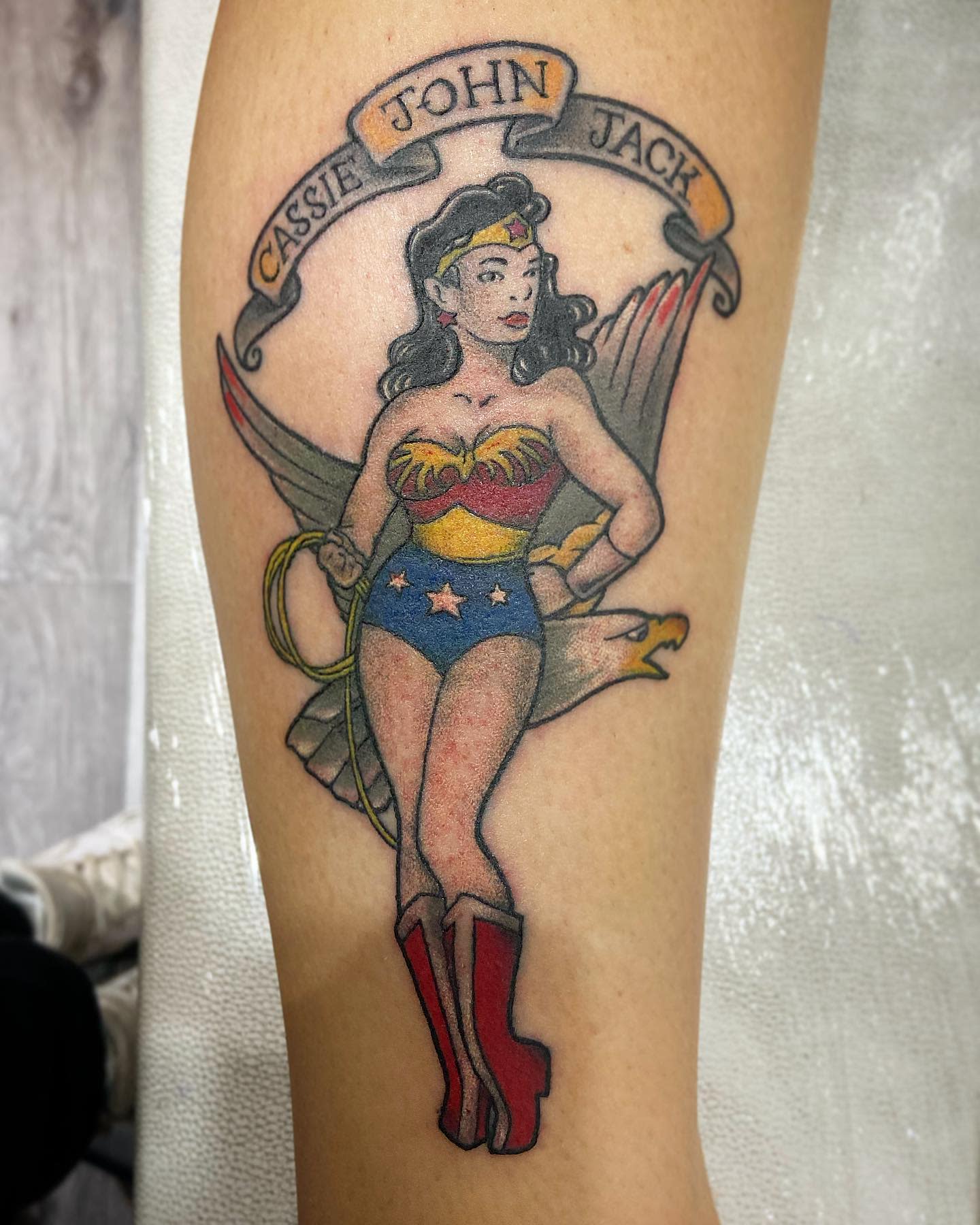 Pin-up style Wonder Woman tattoo with a vintage look, featuring a banner with names above her